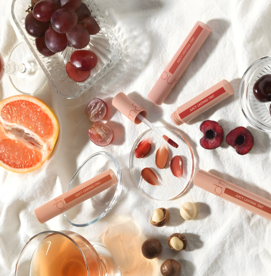 Lip glosses with grapes, cherries, and a glass of juice on a white surface